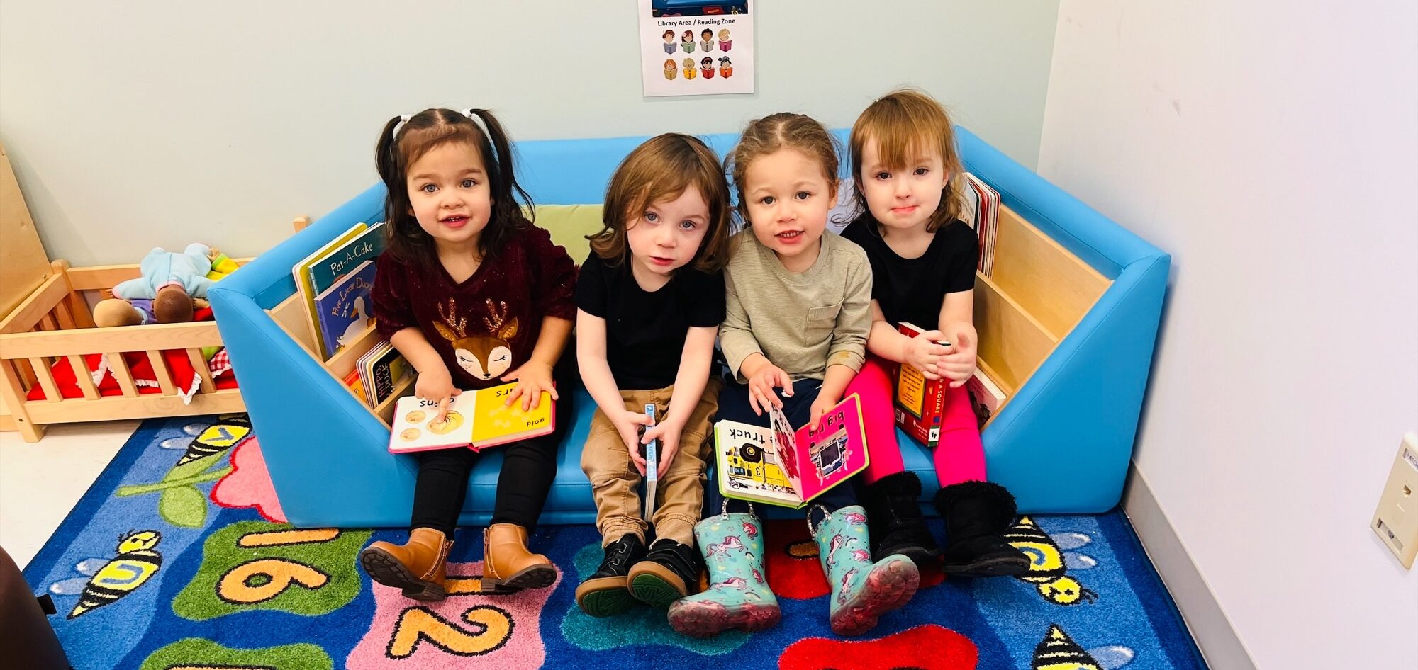 Four toddler students sitting in their classroom reading area and each holding a book.